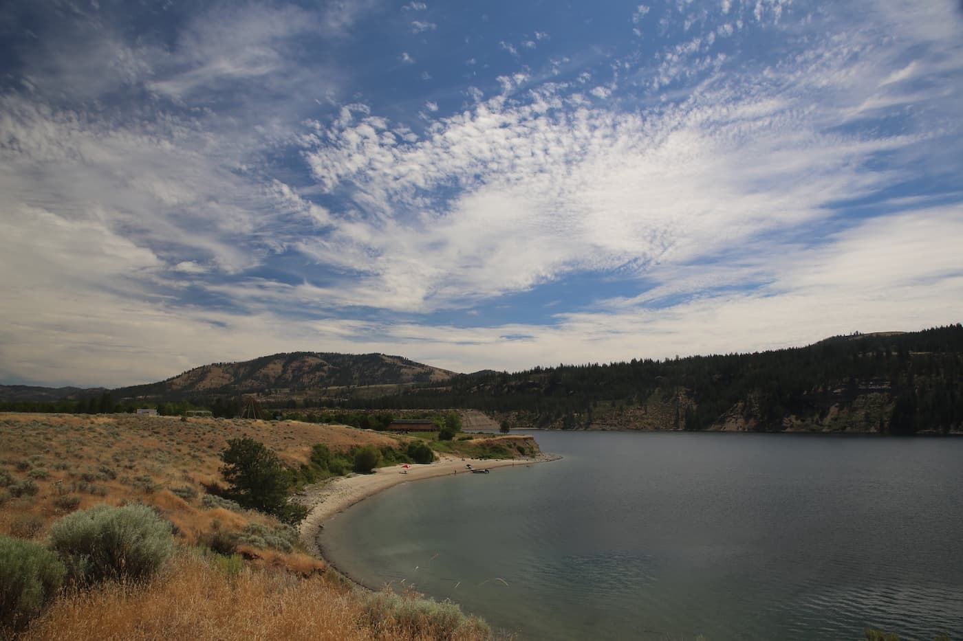 A curved gravel beach on a calm lake, below sagebrush slopes and forested hills, under a wide sky of streaked cloud.
