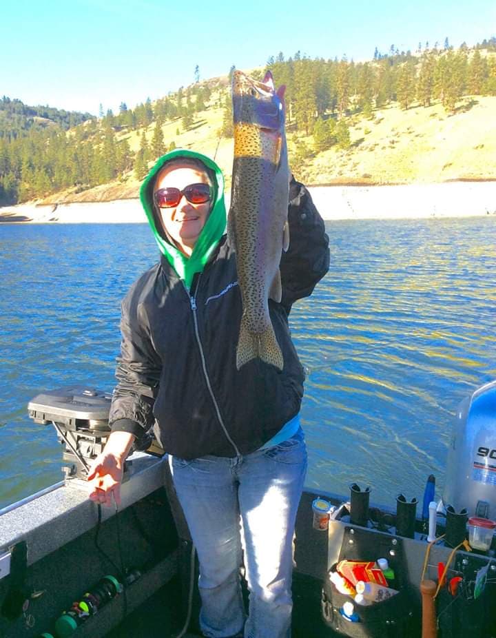 An angler in a green hood and sunglasses holds up a big spotted trout in a boat on the lake, with pine-covered hills behind.